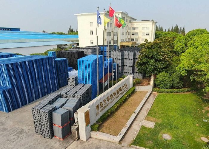 An aerial view of Qingdao Lansu Manufacturing Co., Ltd. The facility has stacks of blue and grey plastic pallets. Several flags are flying on poles in front of a building, surrounded by greenery, indicating a well - maintained manufacturing site.